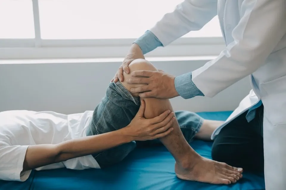 Un medic într-o halat albă examinează genunchiul unui pacient care stă întins pe o masă de examinare albastră.