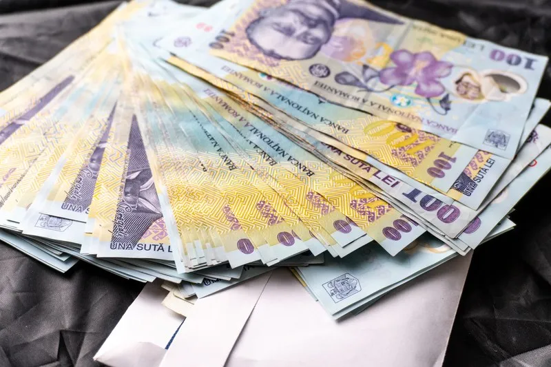 Un teanc de bancnote de o sută de lei este întins pe o suprafață închisă și neuniformă.