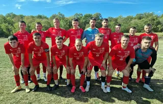 Un grup de bărbați îmbrăcați în echipamente sportive roșii și albastre pozează într-o poziție de echipă pe un teren de fotbal cu iarbă verde.