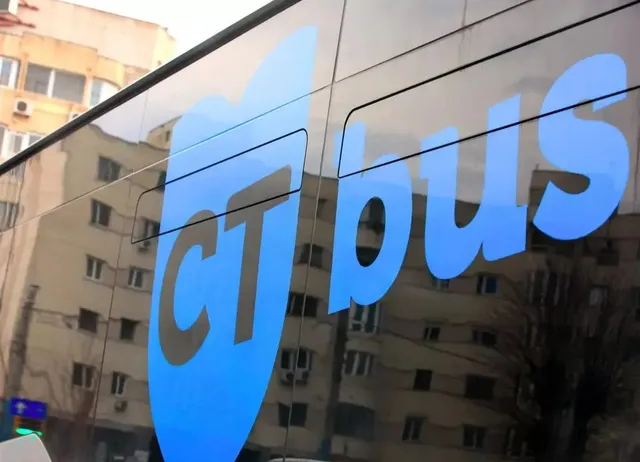 Un logo albastru cu textul "ct bus" este aplicat pe o suprafață de sticlă care reflectă clădiri urbane și arbori.