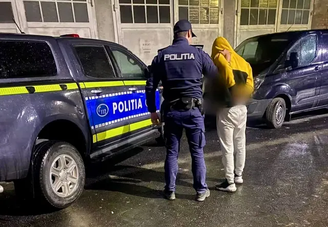 Un polițist în uniformă stă lângă un vehicul de serviciu și un bărbat îmbrăcat în hanorac galben și pantaloni albi.