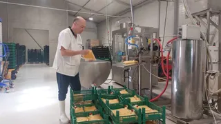 Un bărbat în șapcă, costum alb și cizme albeversează unui container metalic într-o fabrică, în jurul lui fiind cutii verzi cu fructe și echipamente industriale.