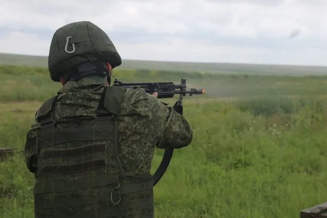 Un soldat în uniformă de camuflaj și vestă tactică țintește un pușcă de la spate într-un câmp deschis sub un cer înnorat.