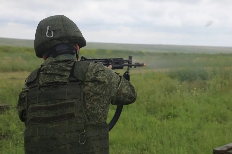 Un soldat în uniformă de camuflaj și vestă tactică țintește un pușcă de la spate într-un câmp deschis sub un cer înnorat.