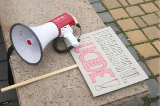 Un megafon alb și roșu cu o curea neagră stă pe o treaptă de piatră lângă un carton alb cu text roz și negru.