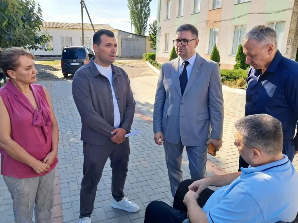 Patru persoane stau în picioare pe o curte pavată lângă o clădire, în timp ce un bărbat stă așezat pe un scaun.