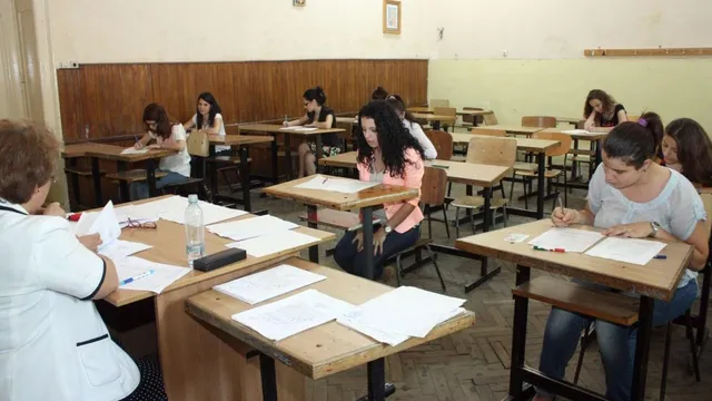 Mai mulți elevi stau la bănci în interiorul unei săli de clasă, scriind pe foi de hârtie în fața unor caiete și a unor sticle de apă.