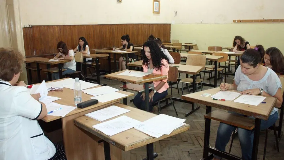 Mai mulți elevi stau la bănci în interiorul unei săli de clasă, scriind pe foi de hârtie în fața unor caiete și a unor sticle de apă.