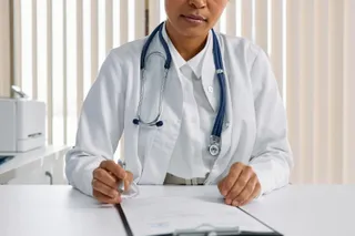 O persoană în halat alb cu un stetoskop la gât scrie pe o foaie de pe un dosar de clinică aflat pe o masă albă.