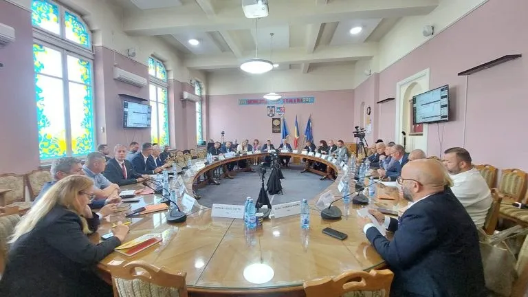 Oamenii stau la mese rotunde de lemn într-o sală cu pereți roz, sub ferestre înalte și un proiector suspendat în tavan.