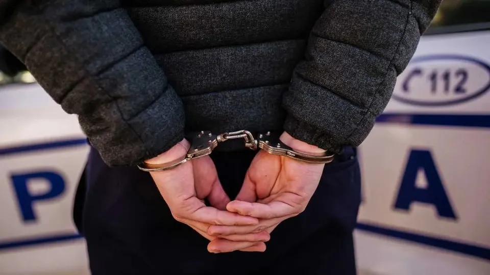 Un bărbat poartă o jachetă neagră și are mâinile în urma spatelui, fiind restricționat cu cătușe metalice în fața unui vehicul alb cu inscripții albastre.