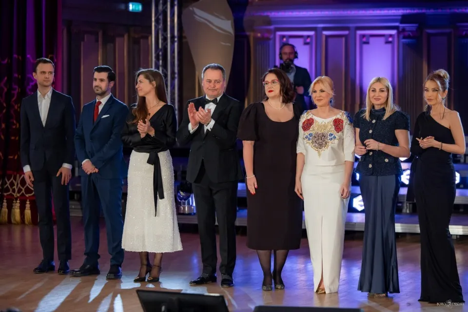 Un grup de oameni în haine de seară stă în rând pe o scenă iluminată cu lumini mov și albastre.