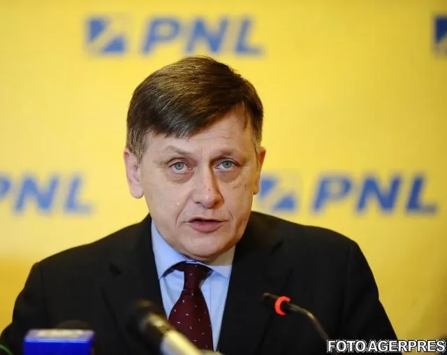 Un bărbat cu părul scurt și haine închise stă în fața unui fundal galben cu inscripții albastre, având microfoane în prim-plan.