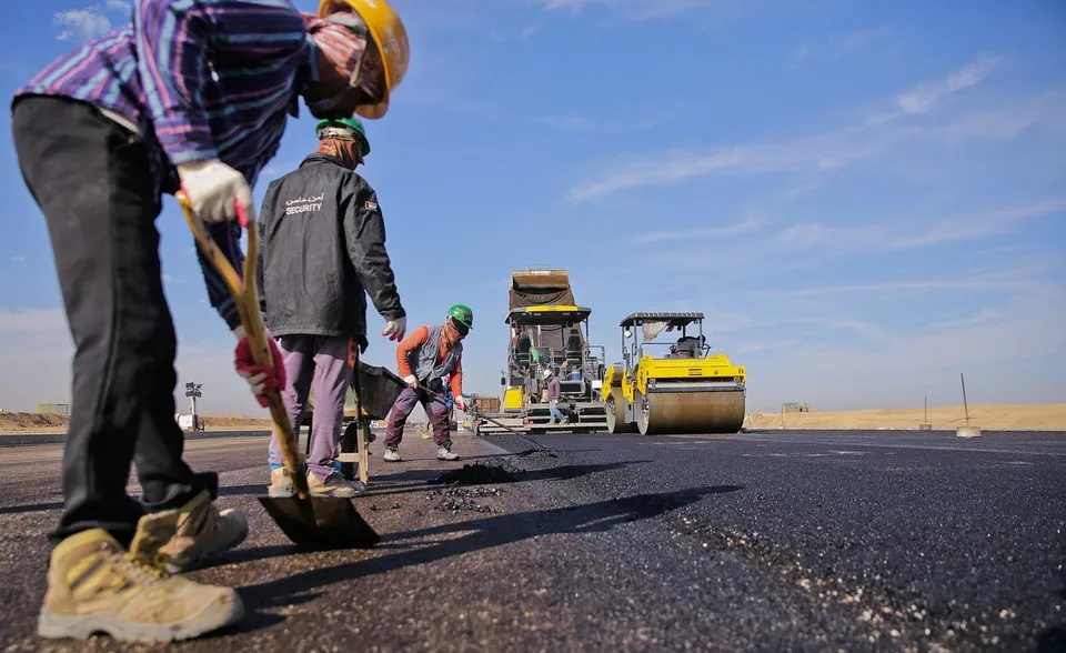 Lucrători în căști și haine de protecție așterne asfalt proaspăt lângă utilaje de construcție, pe un drum larg sub cer senin.