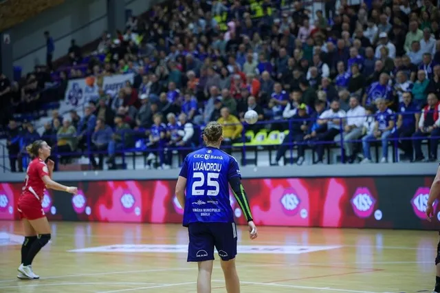 O jucătoare de handbal în echipament roșu și un jucător în echipament albastru cu numărul 25 se află pe un teren de sport în fața tribunelor.