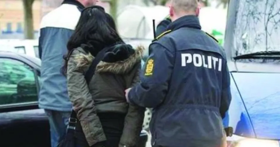 Un polițist în uniformă neagră cu inscripția POLITI pe spate stă lângă o femeie cu părul negru purtând o haină cu blană.