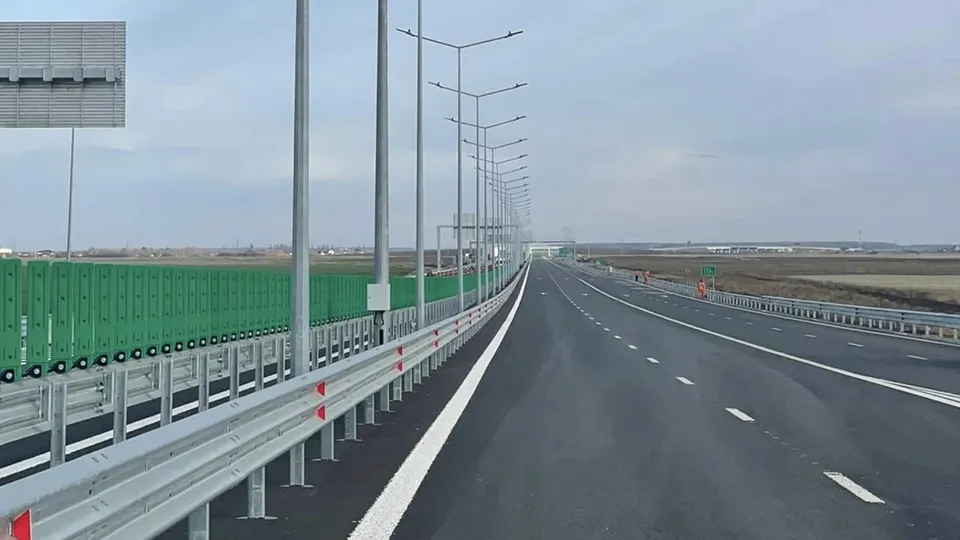 O autostradă cu mai multe benzi de rulare, delimitată de bariere metalice și stâlpi de iluminat pe un fundal de câmp deschis sub un cer înnorat.