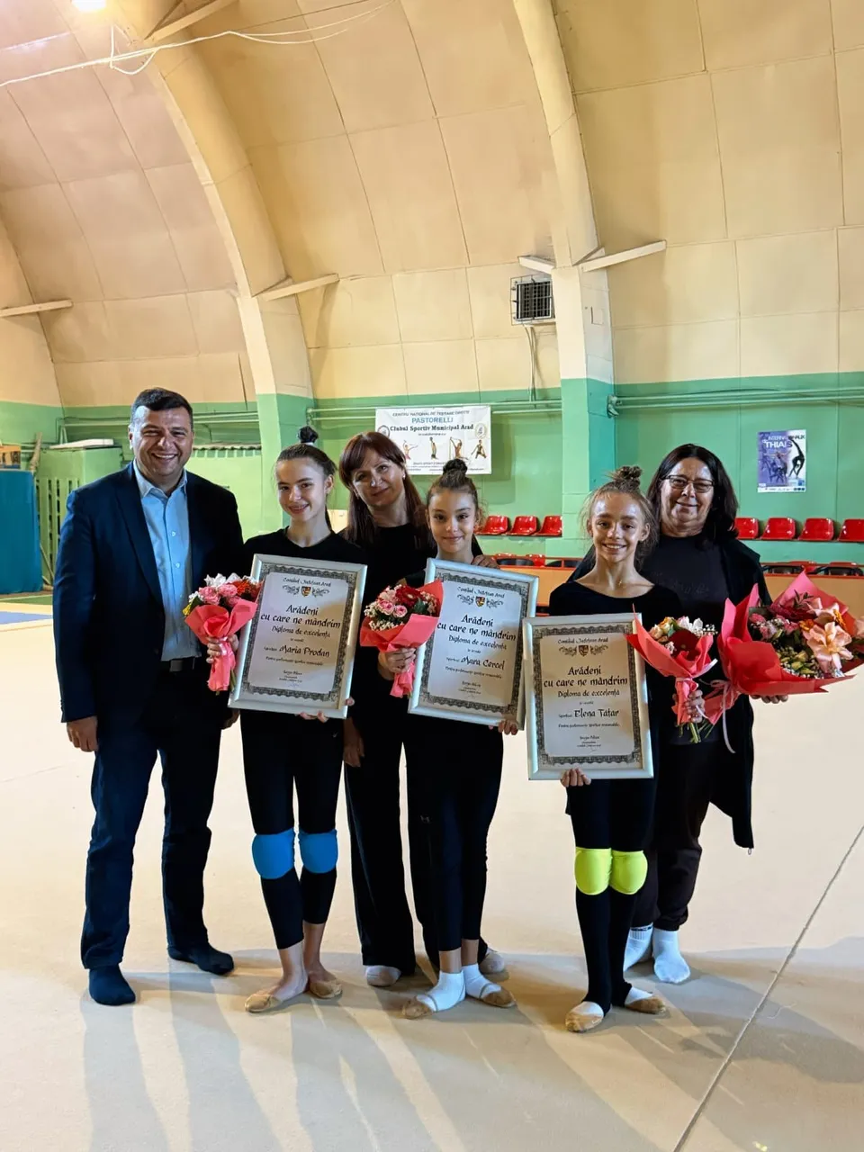 Cinci persoane stau în rând într-o sală de sport, purtând diplome încadrate și buchete de flori roșii.