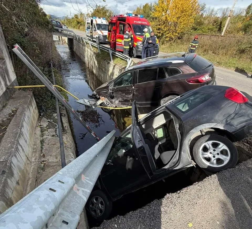 Două mașini negre sunt oprite pe marginea unui șanț, lângă un parapet metalic, în timp ce vehicule de urgență și personal sunt prezente pe drum.