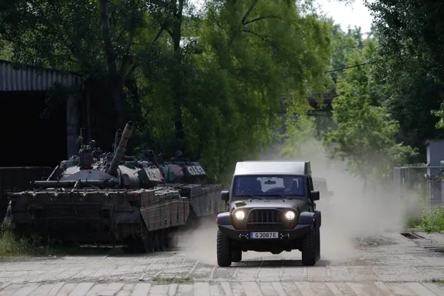 Un vehicul militar de culoare închisă circulă pe un drum de pământ, ridicând un nor de praf lângă câteva tancuri parcate sub copaci.