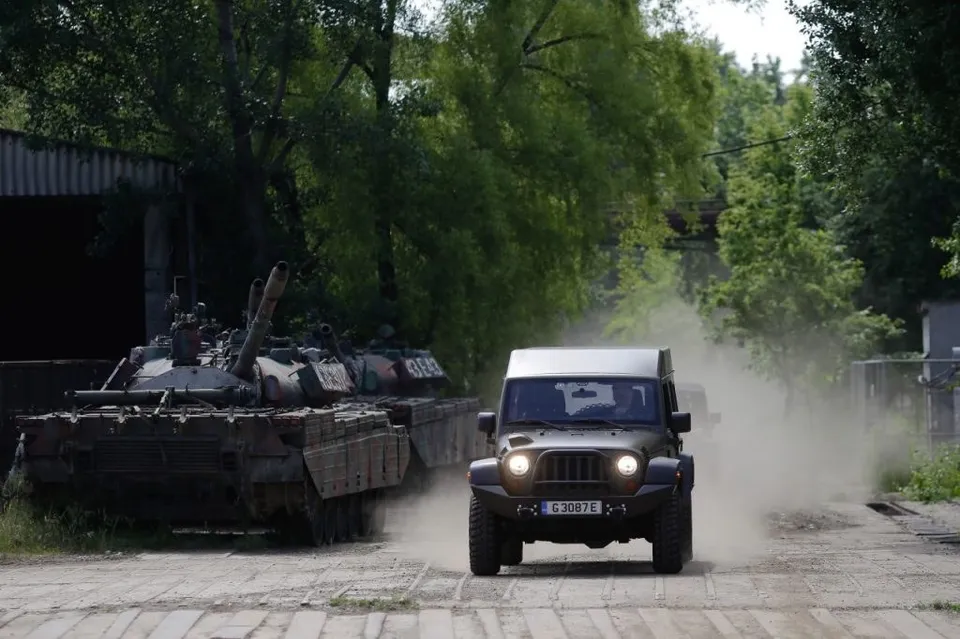 Un vehicul militar de culoare închisă circulă pe un drum de pământ, ridicând un nor de praf lângă câteva tancuri parcate sub copaci.
