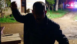 Un bărbat cu gura deschisă și mâna dreaptă ridicată stă într-un spațiu exterior întunecat lângă o masă de lemn.