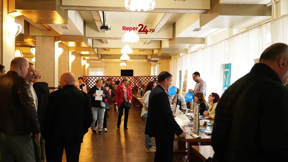 Oamenii stau în picioare într-o încăpere interioară lângă mese pe care sunt așezate diverse obiecte și documente.