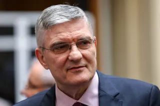 Un bărbat cu părul cărunt și ochelari poartă o cămașă roz și un sacou închis la culoare.