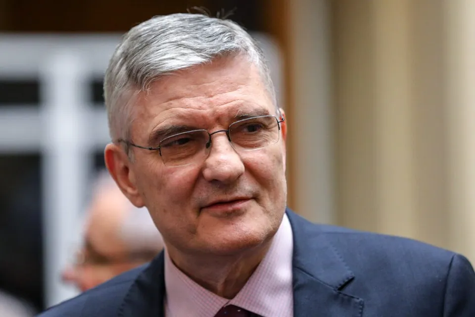 Un bărbat cu părul cărunt și ochelari poartă o cămașă roz și un sacou închis la culoare.