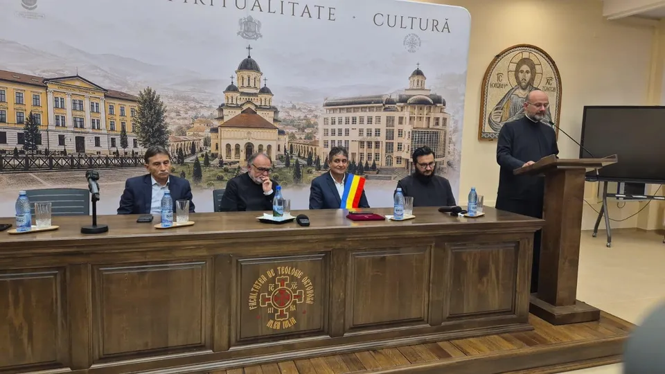 Cinci bărbați stau în spatele unei mese lungi de lemn într-o încăpere cu un panou decorativ în fundal și un icon în partea dreaptă.