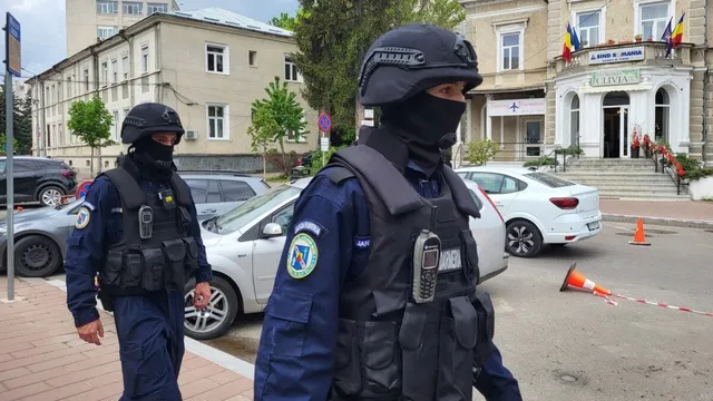Doi polițiști în uniformă albastră și vestă tactică neagră cu mască facială merg pe un trotuar lângă mașini parcate.