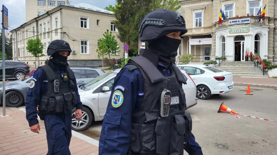 Doi polițiști în uniformă albastră și vestă tactică neagră cu mască facială merg pe un trotuar lângă mașini parcate.