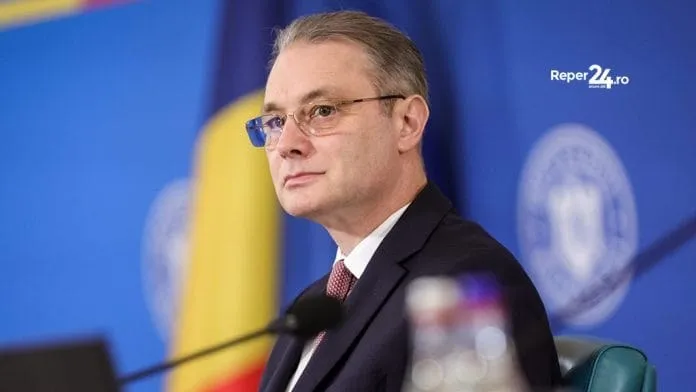 Un bărbat cu ochelari și costum închis la culoare stă în fața unui fundal albastru cu elemente de steag și embleme circulare.