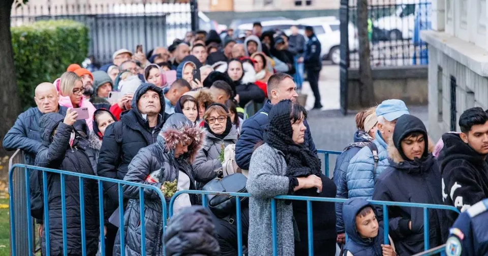 O mulțime de oameni îmbrăcați în haine de iarnă stă în spatele unor garduri metalice albastre într-un spațiu exterior.