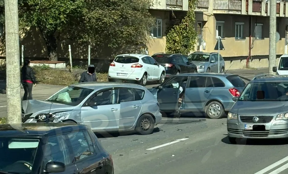 Două autoturisme de culoare gri sunt implicate într-o coliziune pe un drum, în timp ce alte vehicule și persoane sunt vizibile în proximitate.
