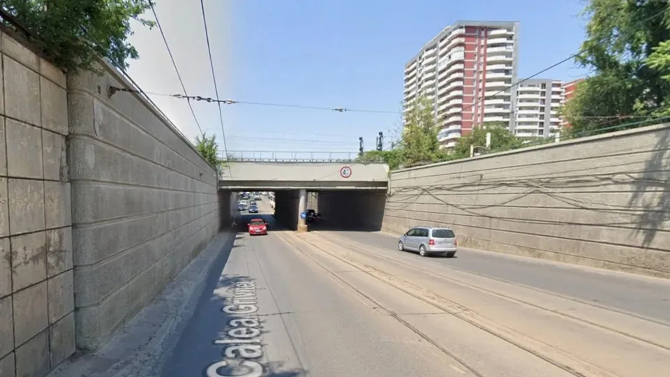 O stradă asfaltată cu șine de tramvai trece pe sub un pod de beton, flancată de ziduri înalte și clădiri înalte în fundal.