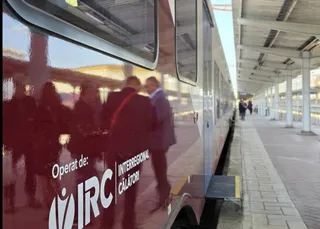 Un tren roșu stă pe un peron de gară lângă un acoperiș alb, cu siluete de persoane reflectate pe fereastra laterală a vagonului.