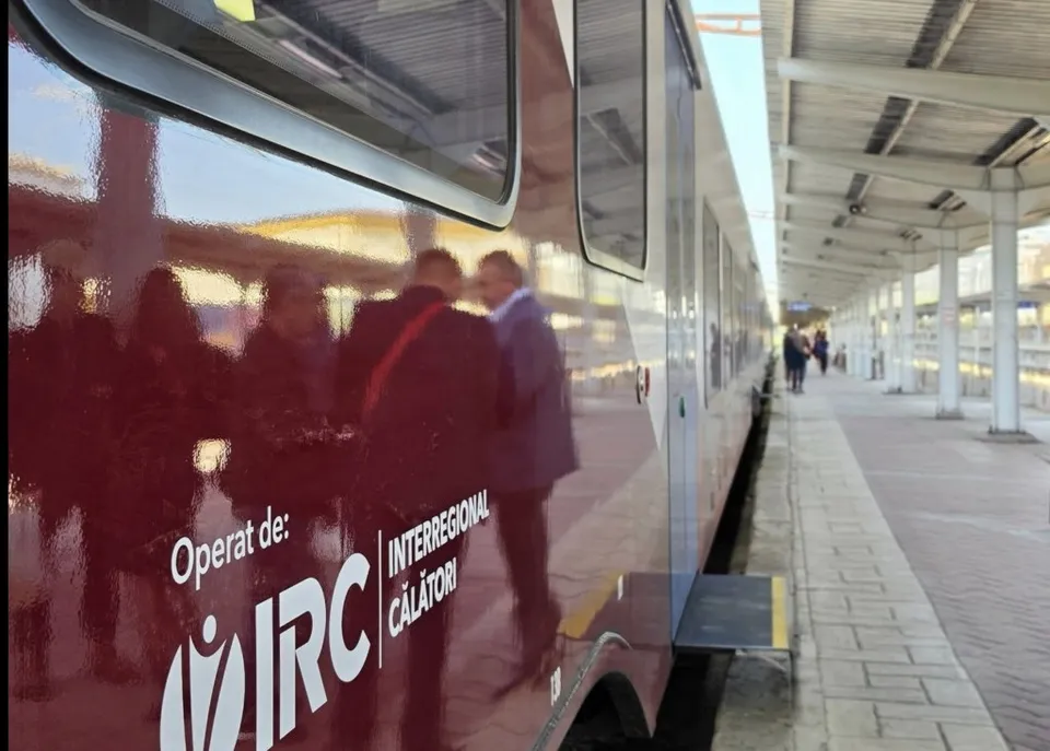 Un tren roșu stă pe un peron de gară lângă un acoperiș alb, cu siluete de persoane reflectate pe fereastra laterală a vagonului.