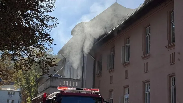 Fum alb gros se ridică de deasupra acoperișului unei clădiri de culoare roz lângă un vehicul de serviciu cu lumini roșii aprinse.