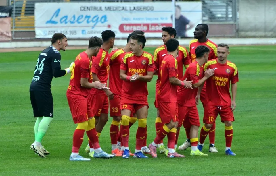 Un grup de jucători de fotbal în echipamente roșii și unul în echipament negru și verde stau pe un teren de fotbal.