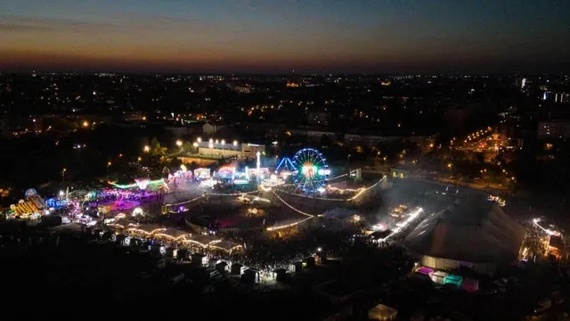 O vedere aeriană nocturnă asupra unui parc de distracții iluminat cu diverse structuri, un turn de observare rotativ și lumină difuză pe fundalul unui oraș.