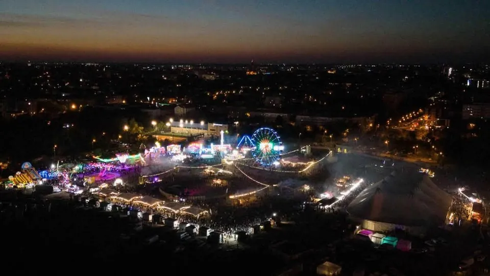 O vedere aeriană nocturnă asupra unui parc de distracții iluminat cu diverse structuri, un turn de observare rotativ și lumină difuză pe fundalul unui oraș.