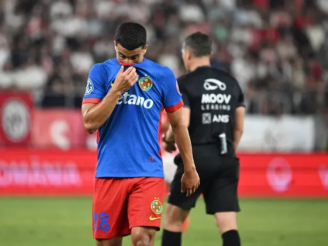 Un fotbalist într-un echipament albastru și roșu stă pe teren, cu privirea în jos, lângă un arbitru care poartă tricou negru.