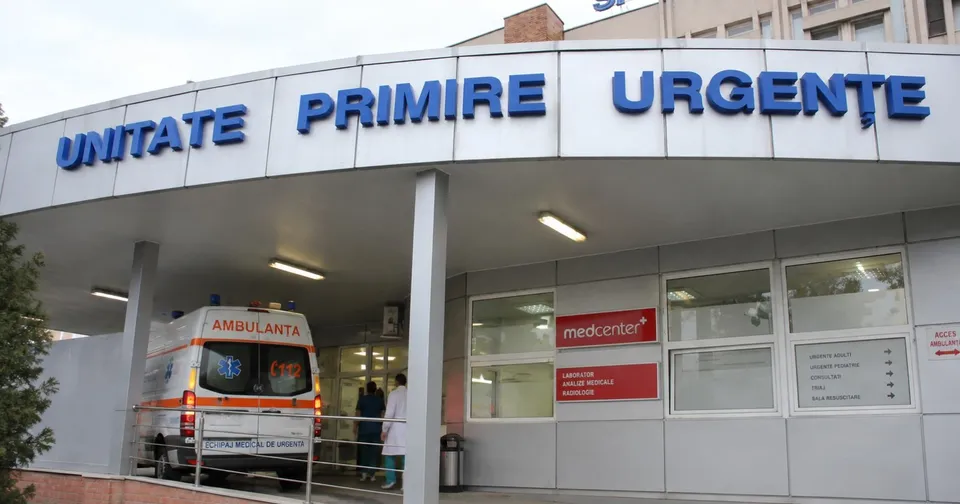 O ambulanță albă cu inscripții roșii este parcată sub un acoperiș de clădire pe care scrie UNITATE PRIMIRE URGENȚE în litere albastre.