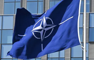 Un steag albastru cu simbolul alb al NATO flutură pe un stârf în fața unei clădiri cu ferestre mari de sticlă.
