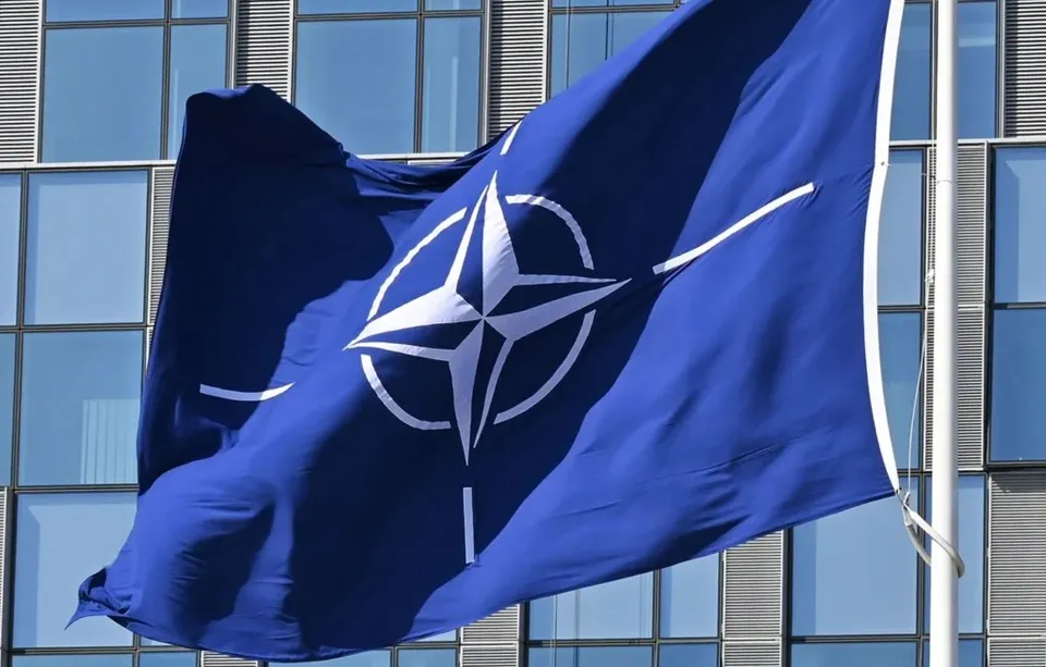 Un steag albastru cu simbolul NATO este agitat pe un stârf în fața unei clădiri cu ferestre mari de sticlă.