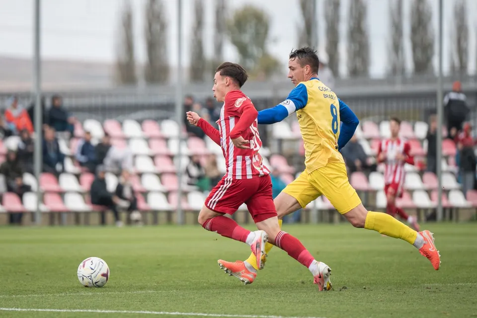 Un jucător în echipament roșu și alb chạy după o minge pe un teren de fotbal, fiind urmărit de un jucător în echipament galben și albastru.