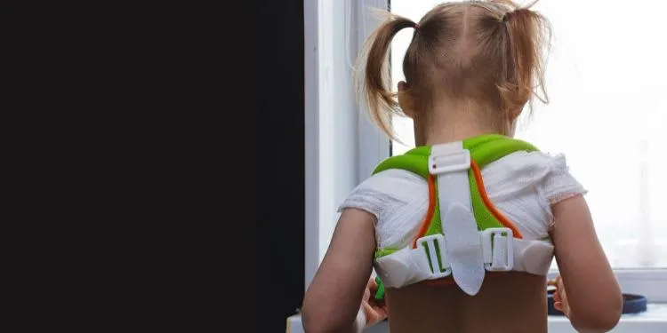 Un copil cu părul blond în cozi poartă un dispozitiv ortopedic de culoare verde și alb pe spate, stând cu spatele la cameră în fața unei ferestre.