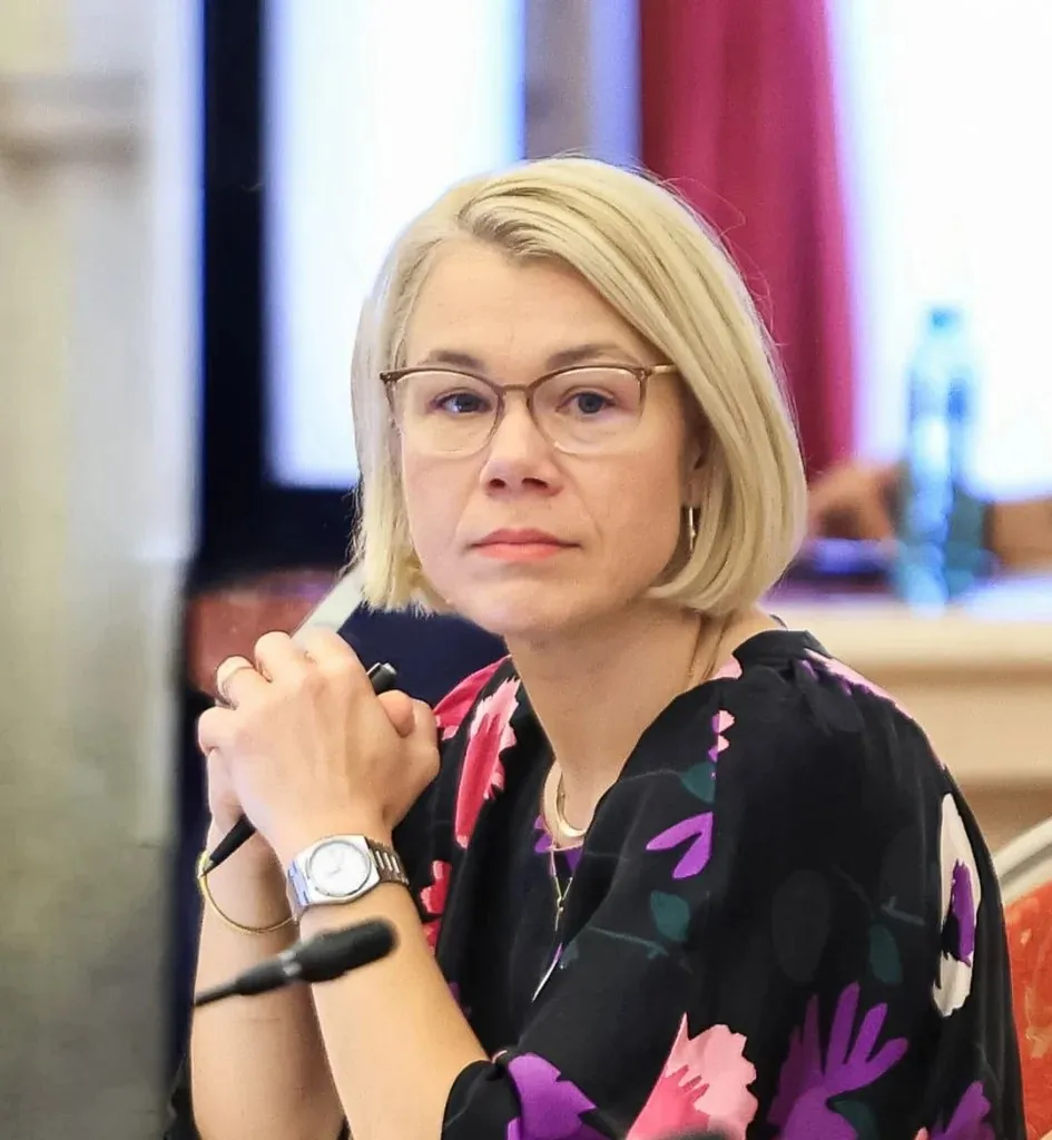 O femeie cu părul blond și ochelari, purtând o bluză neagră cu motive florale, stă cu mâinile împreună lângă un microfon.