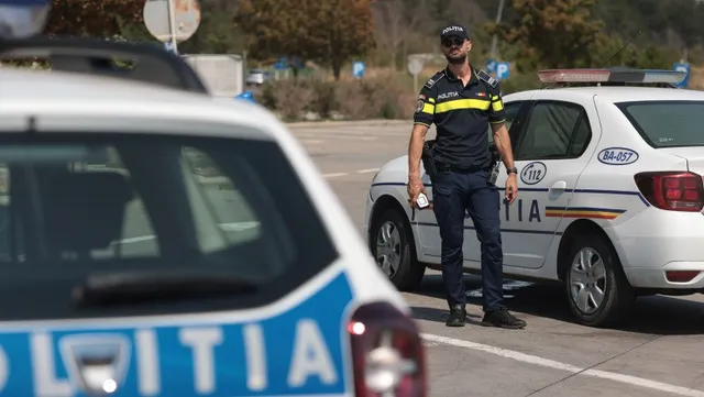 Un polițist stă în picioare lângă o mașină de poliție albă pe un drum asfaltat.